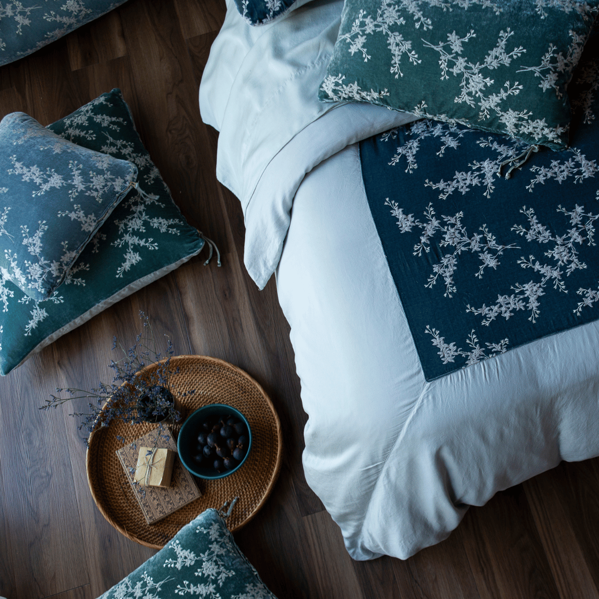 : an overhead shot of a blue duvet cover with green and blue pillows scattered about the bed and dark wood floor.