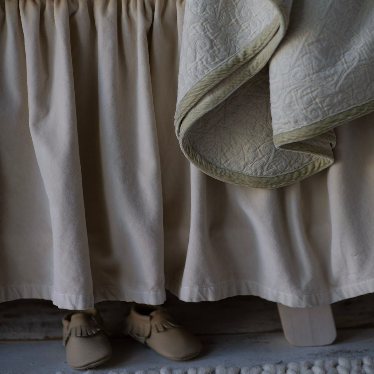 : cotton velvet crib skirt in a neutral tone shown close up with the corner of a draped blanket visible along with tan-colored booties on a white-washed floor.