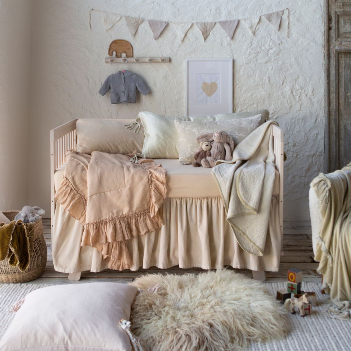 : a neutral crib styled with neutral tones in a nursery scene with adornments on the wall and toys and stuffed animals placed throuughout the scene.