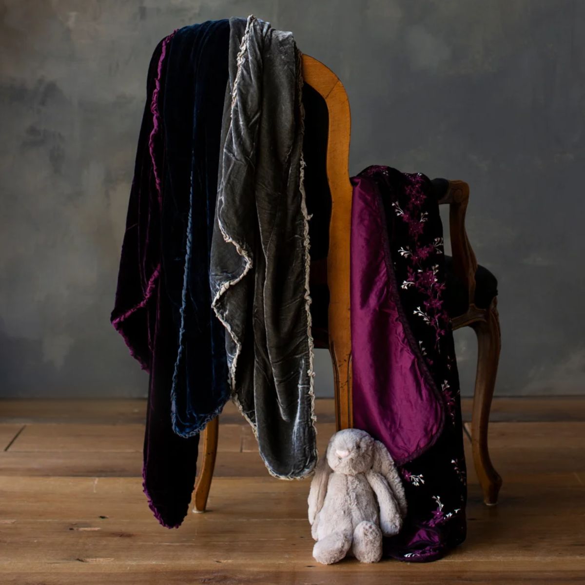 : silk velvet baby blankets in deep jewel tones draped on the back and arm of a chair with a small stuffed bunny on a medium wood floor.