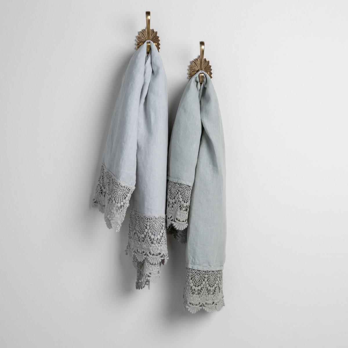 : cloud and eucalyptus linen guest towels with mattine lace hanging from decorative towel hooks against a plain white wall.