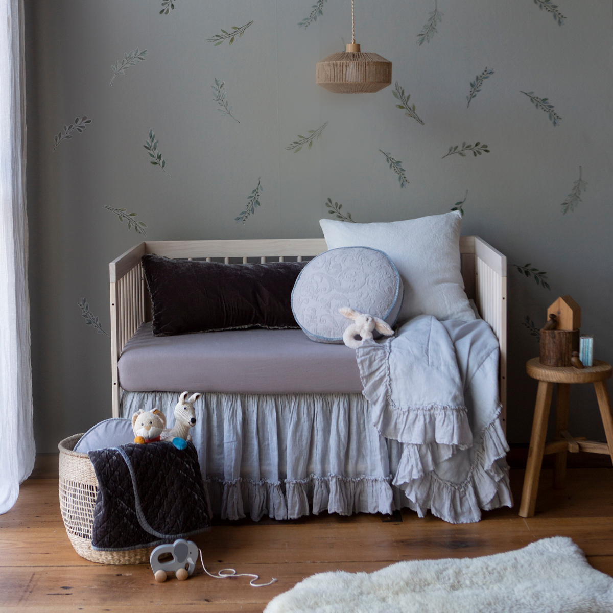 : Blanket in cloud, draped over the front of a crib in blue and grey tones, in nursery with warm wood tones and botanical tone on tone wall paper.