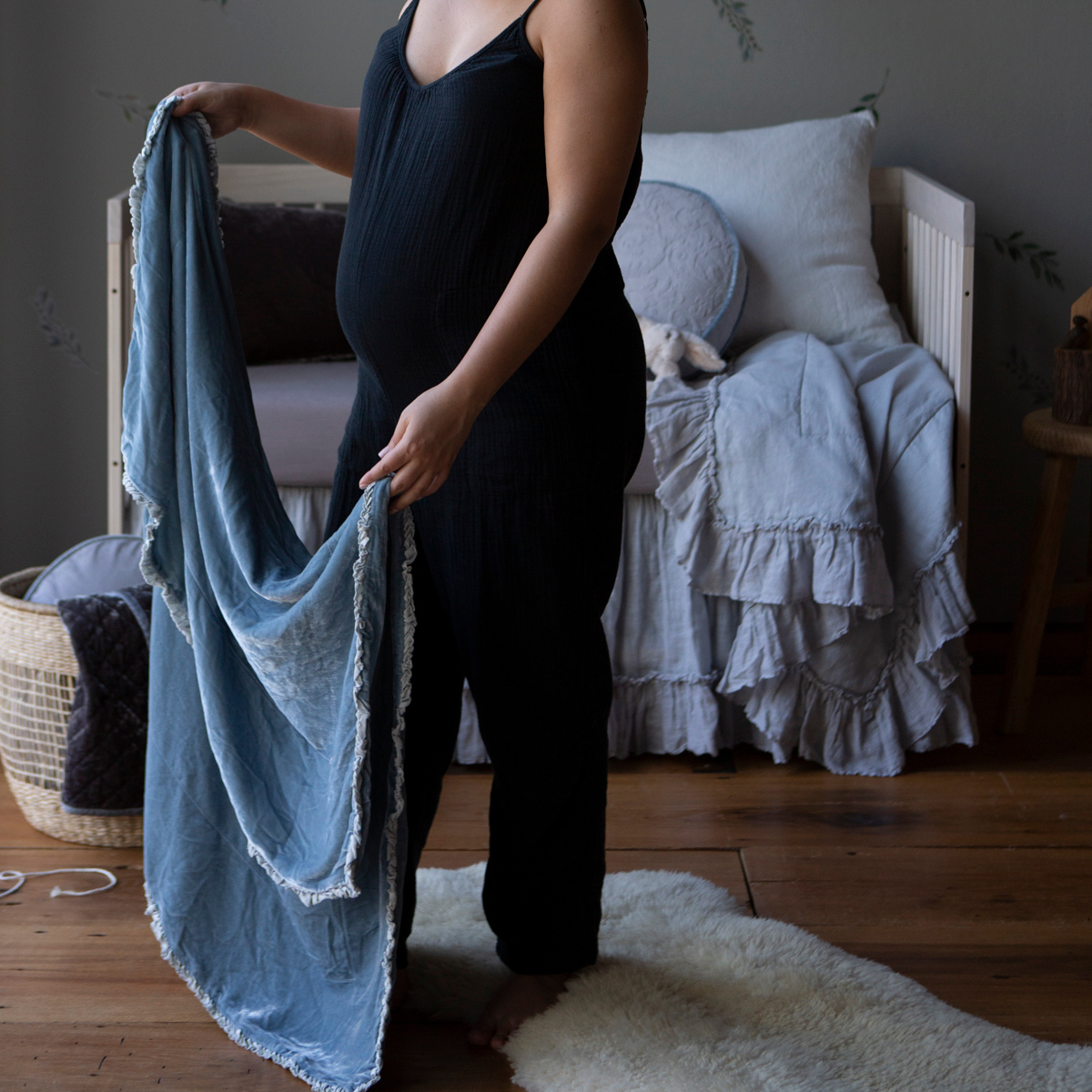 : A pregnant woman, seen from the shoulders down, holding a blanket open in cloud in a blue and grey toned nursery.