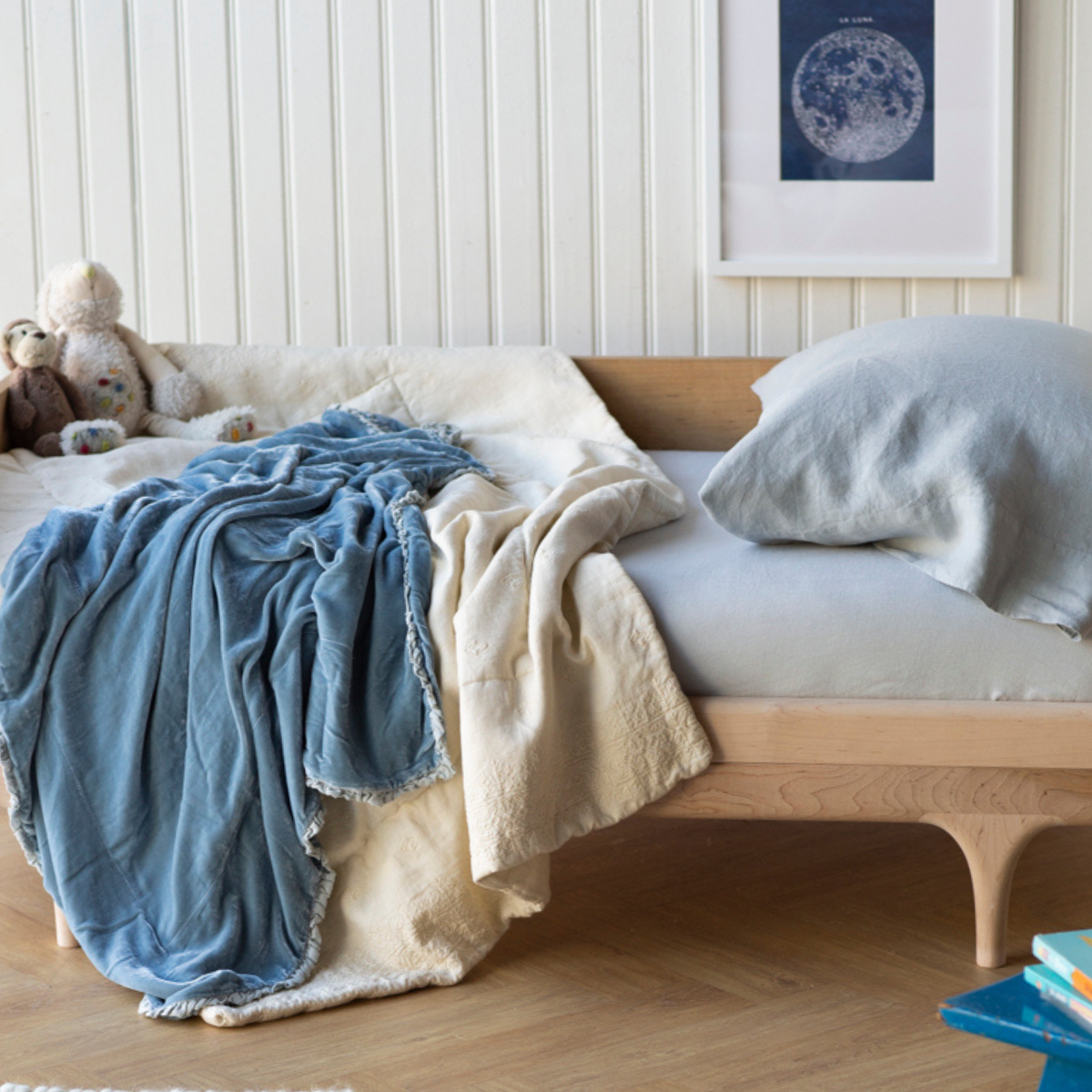 : Blanket in parchment rumpled on a toddler bed, layered with blue and grey bedding in a bright room with warm wood accents.