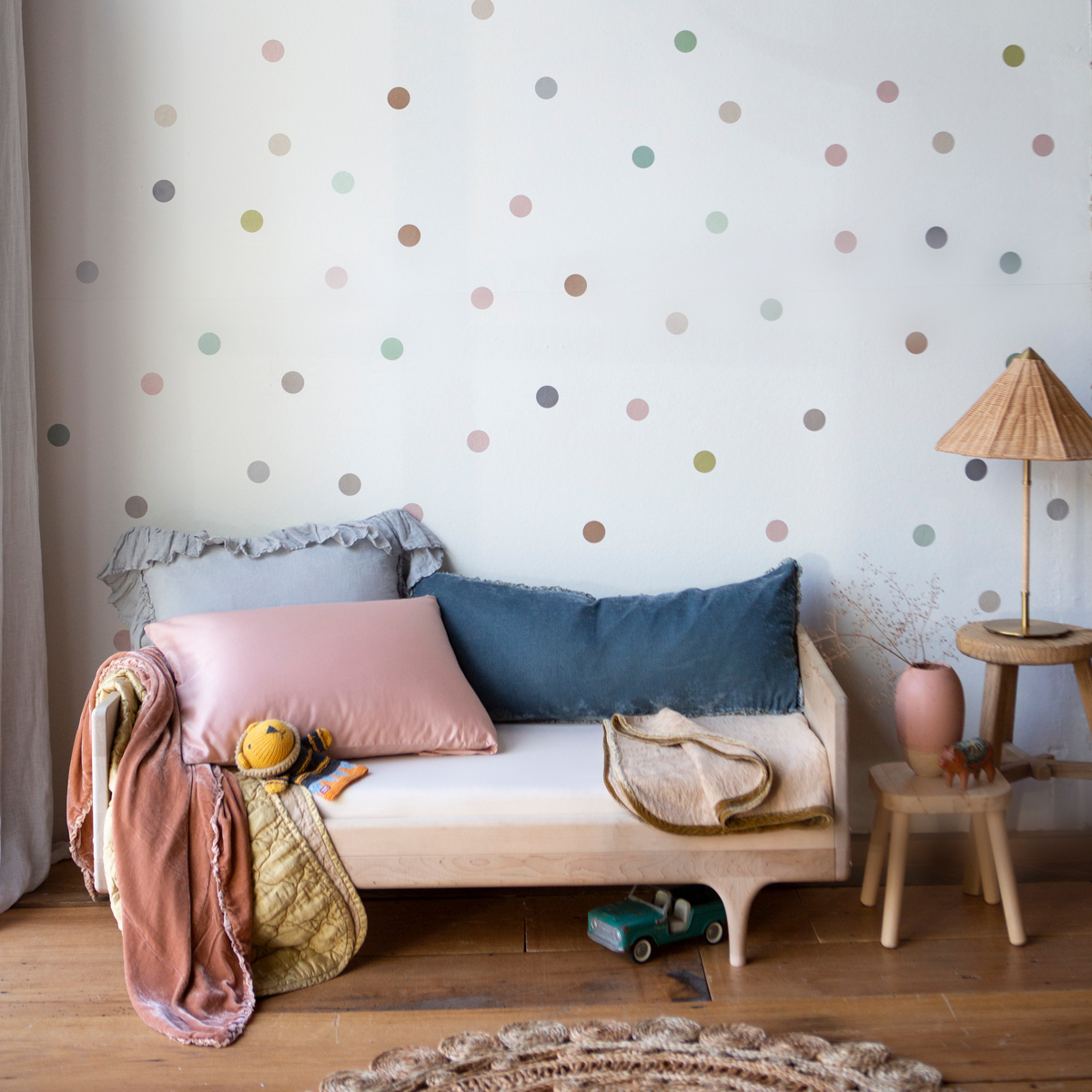 : Crib sheet in parchment on a toddler bed with colorful pillows and blankets in a bright room with warm wood accents.