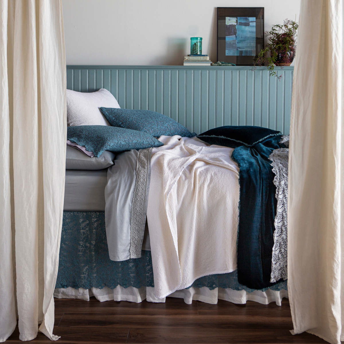 : side view of a bed dressed with lace pillowcases, flat sheet, bed skirt and bed scarf layered with cotton sateen sheets, chenille and cotton jacquard bedspread and shams and silk velvet accent pillow and bed end blanket β shown from the side with curtain panels framing the bed that's against a light blue paneled wall.