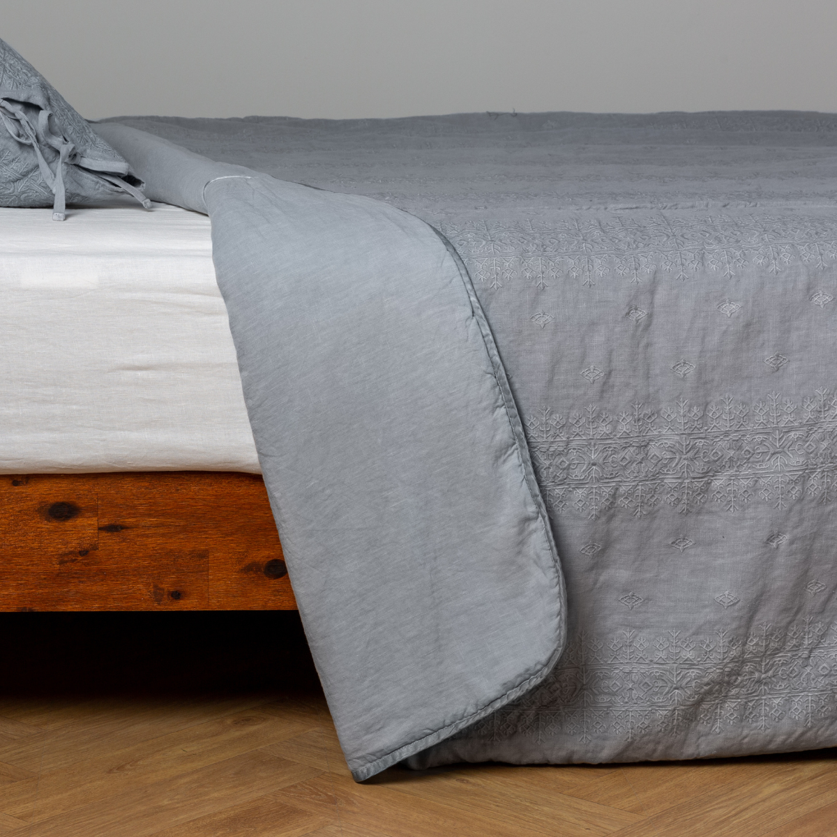 Mineral: White: embroidered bedspread with corner pulled back showing the plain linen back. Shown with a white fitted sheet and matching sham, the bedframe is slightly visible and the bed is against a white wall and on a medium wood floor.
