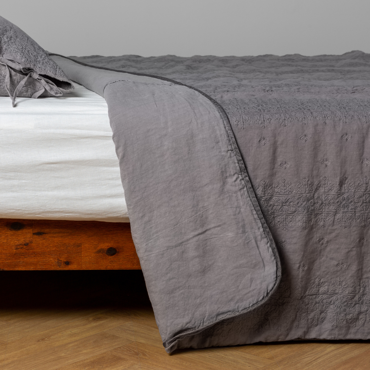 Moonlight: White: embroidered bedspread with corner pulled back showing the plain linen back. Shown with a white fitted sheet and matching sham, the bedframe is slightly visible and the bed is against a white wall and on a medium wood floor.