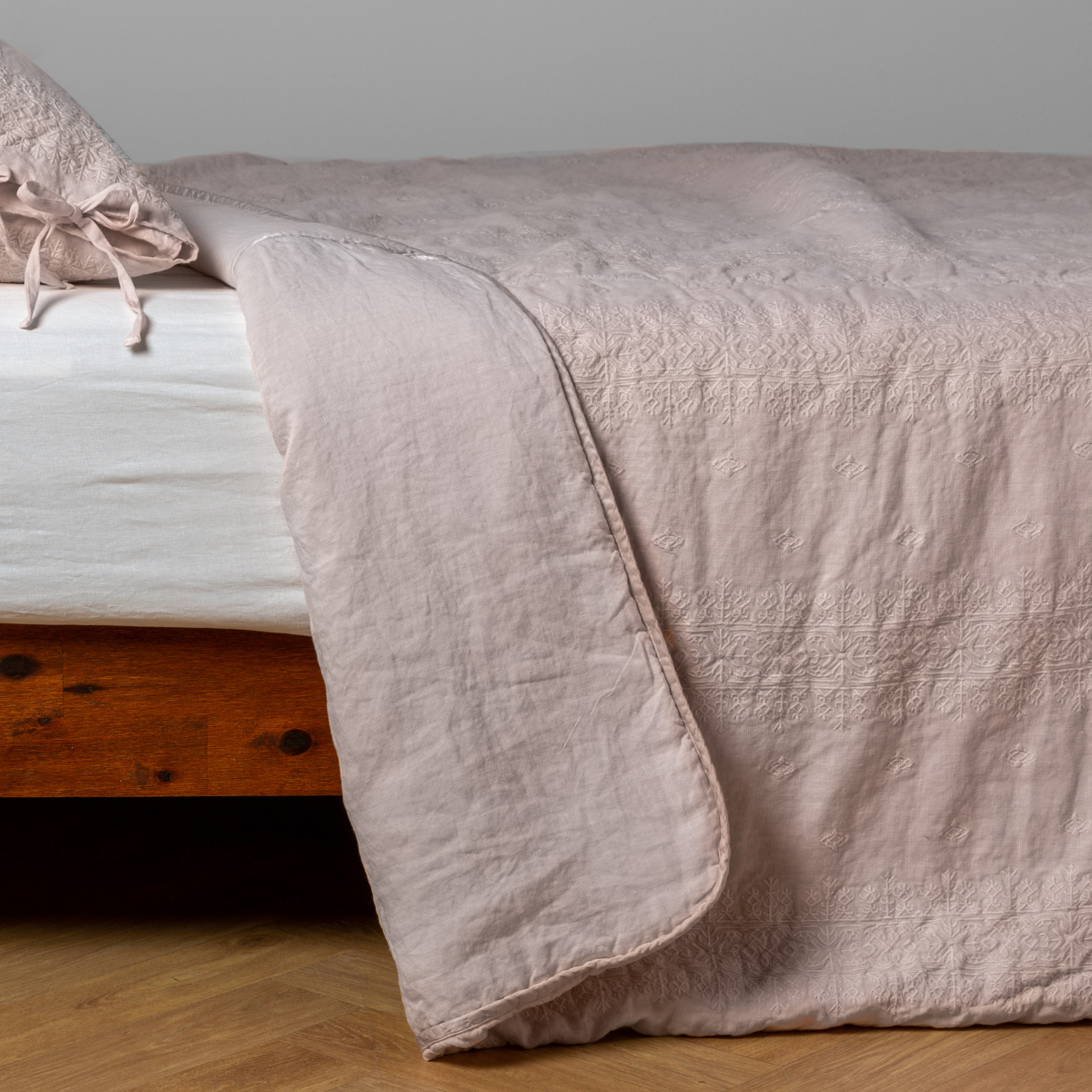 Pearl: White: embroidered bedspread with corner pulled back showing the plain linen back. Shown with a white fitted sheet and matching sham, the bedframe is slightly visible and the bed is against a white wall and on a medium wood floor.