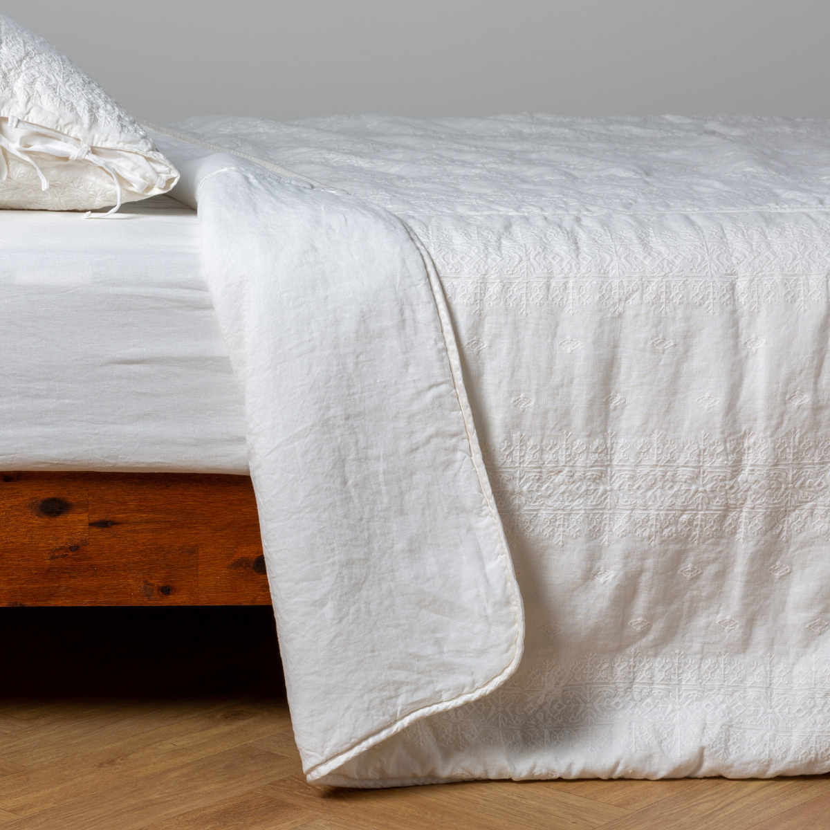 Winter White: White: embroidered bedspread with corner pulled back showing the plain linen back. Shown with a white fitted sheet and matching sham, the bedframe is slightly visible and the bed is against a white wall and on a medium wood floor.
