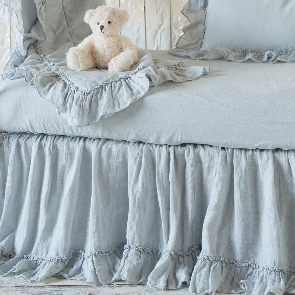 : Linen crib sheet in cloud with matching crib skirt, baby blanket, and throw pillow on a white iron crib decorated with stuffed bear - side view.