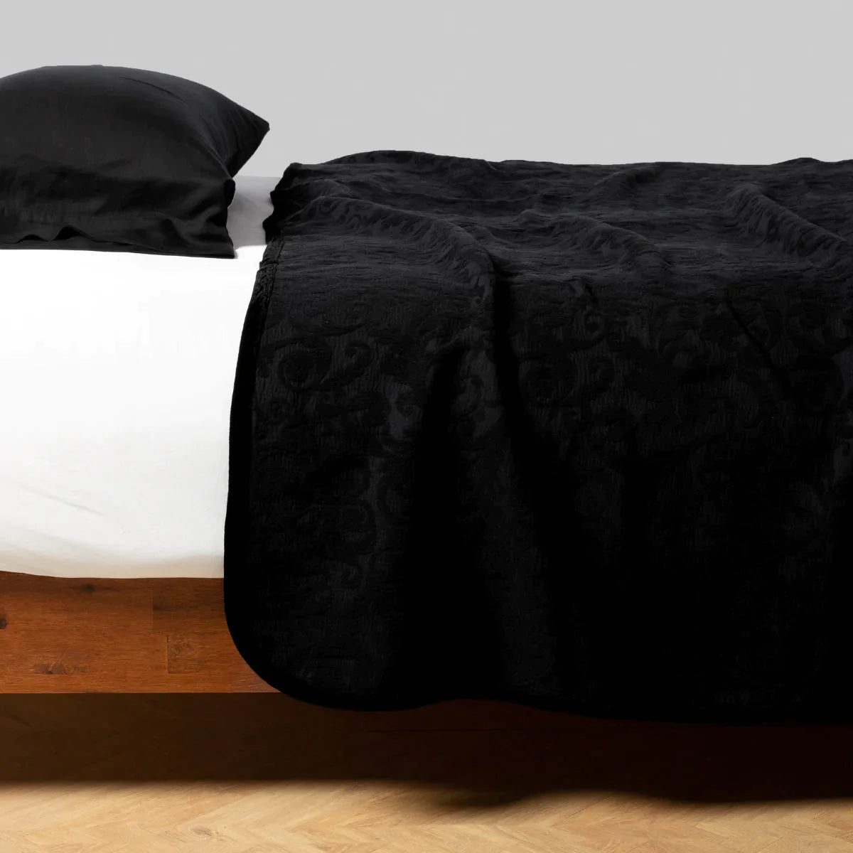 Corvino: The coverlet on a bed viewed from the side against a plain white wall. The bed is styled simply with the coverlet slightly rumpled atop a white sheet. with a pillowcase in Corvino.