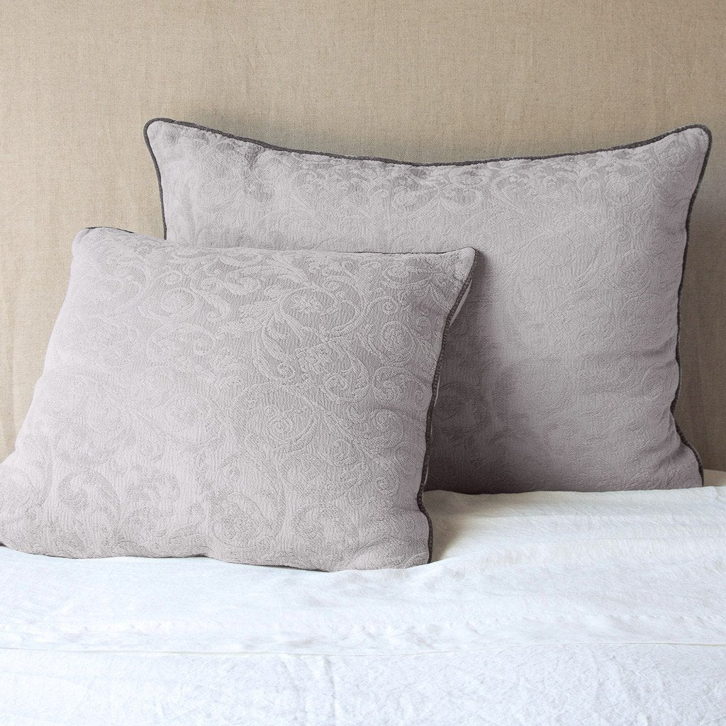 Fog: Two Adele shams, shown in euro and deluxe sizes, leaning against a neutral toned headboard on white bedding.