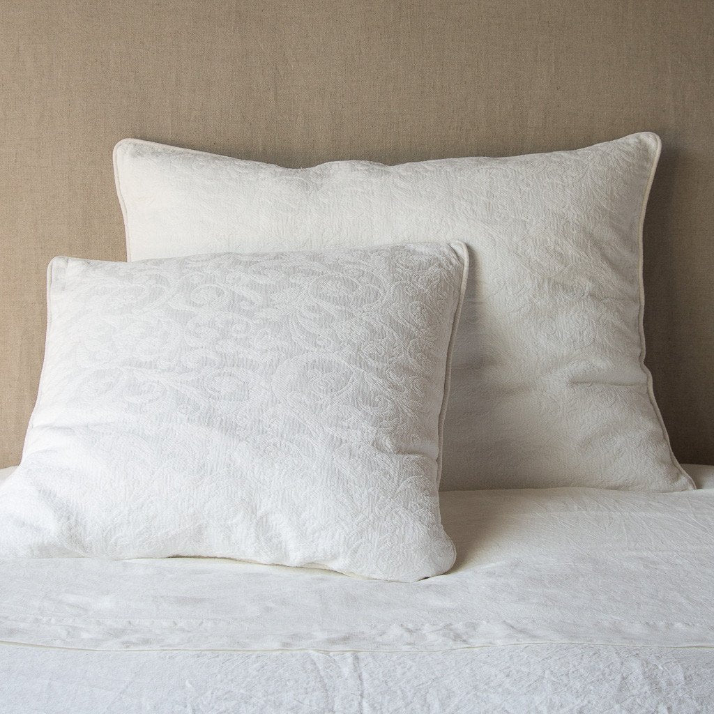 Winter White: Two shams , shown in euro and deluxe sizes, leaning against a neutral toned headboard on white bedding.