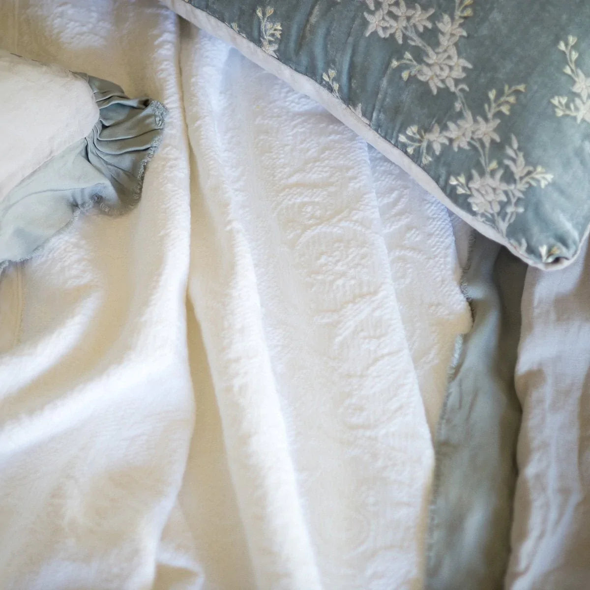 : An overhead close up of a rumpled Adele coverlet in white, surrounded by sterling accent pillows and throw blanket.