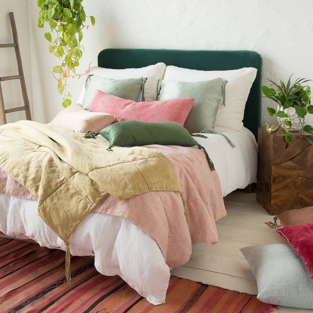 : Shimmery Taline throw blanket and pillows in green and gold tones are paired with pink embroidered linen, with colorful rug, green plants, and warm wood accents - three-quarter angle.
