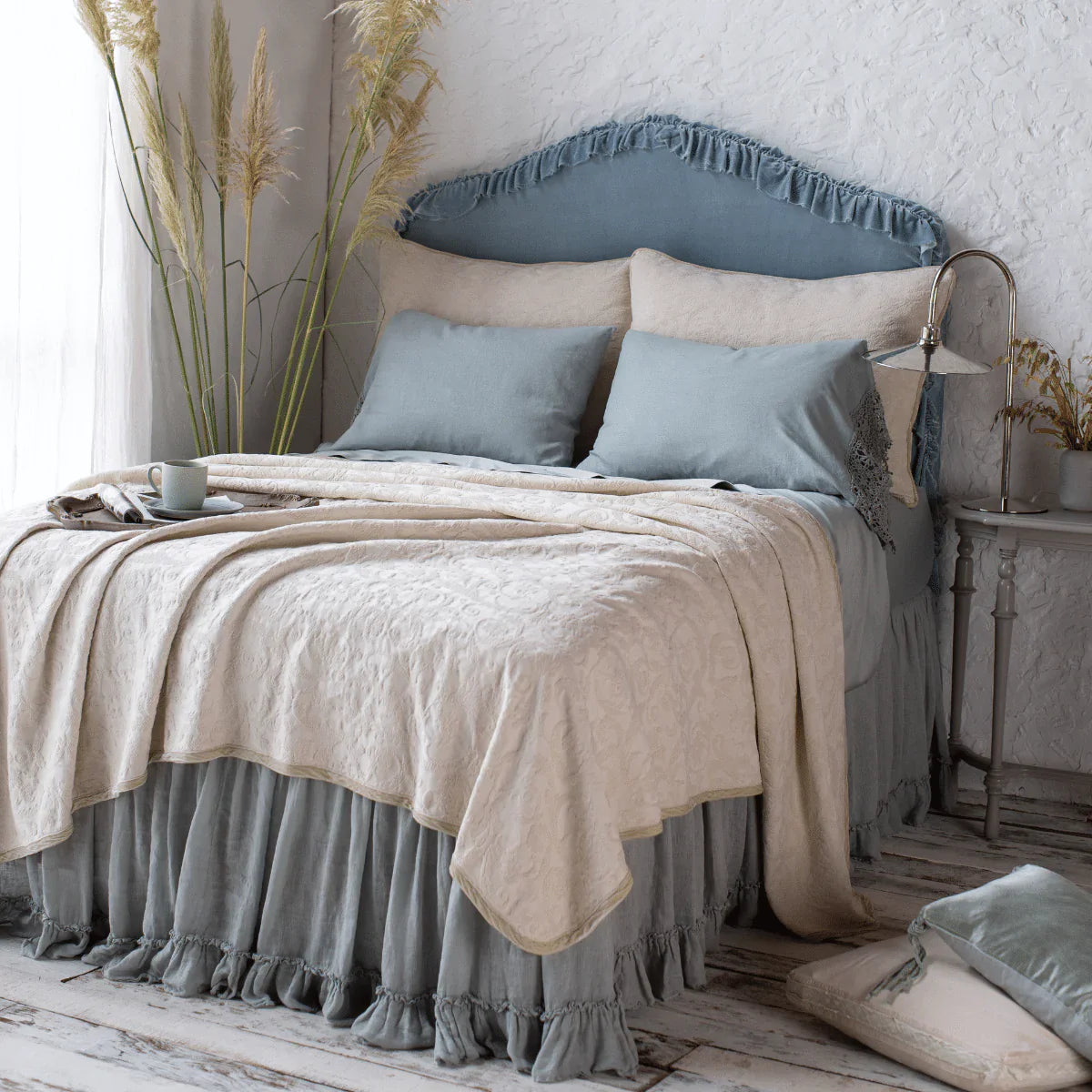 : The Adele coverlet in parchment, shown with matching shams and Bria sheets in eucalyptus. Shot at an angle from the foot of the bed, the coverlet is beautifully rumpled over a ruffled bed skirt. The room in styled on distressed wood floors with dried greenery and natural light.