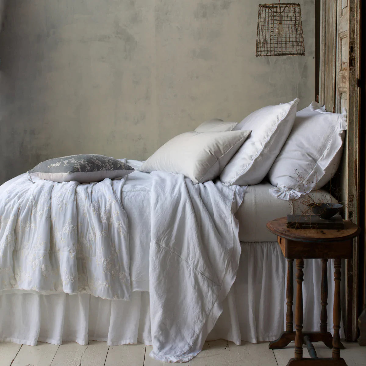 : Sterling Bria sheets contrast against a mostly white bed, shown from the side. The lighting is moody and colors are muted, emphasizing the variety of cotton, linen, and silk textures.