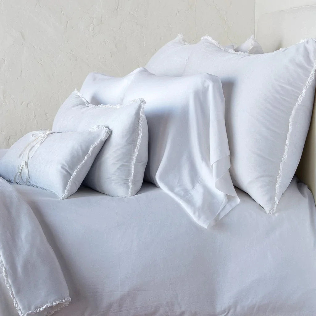 White: Silk velvet shams with a petite ruffle on monochromatic bed, leaning upright behind sleeping and throw pillows - side view.