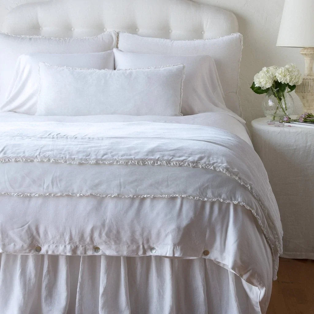 White: Silk velvet shams with a petite ruffle on a monochromatic bed, stacked behind sleeping and throw pillows - view from end of bed.
