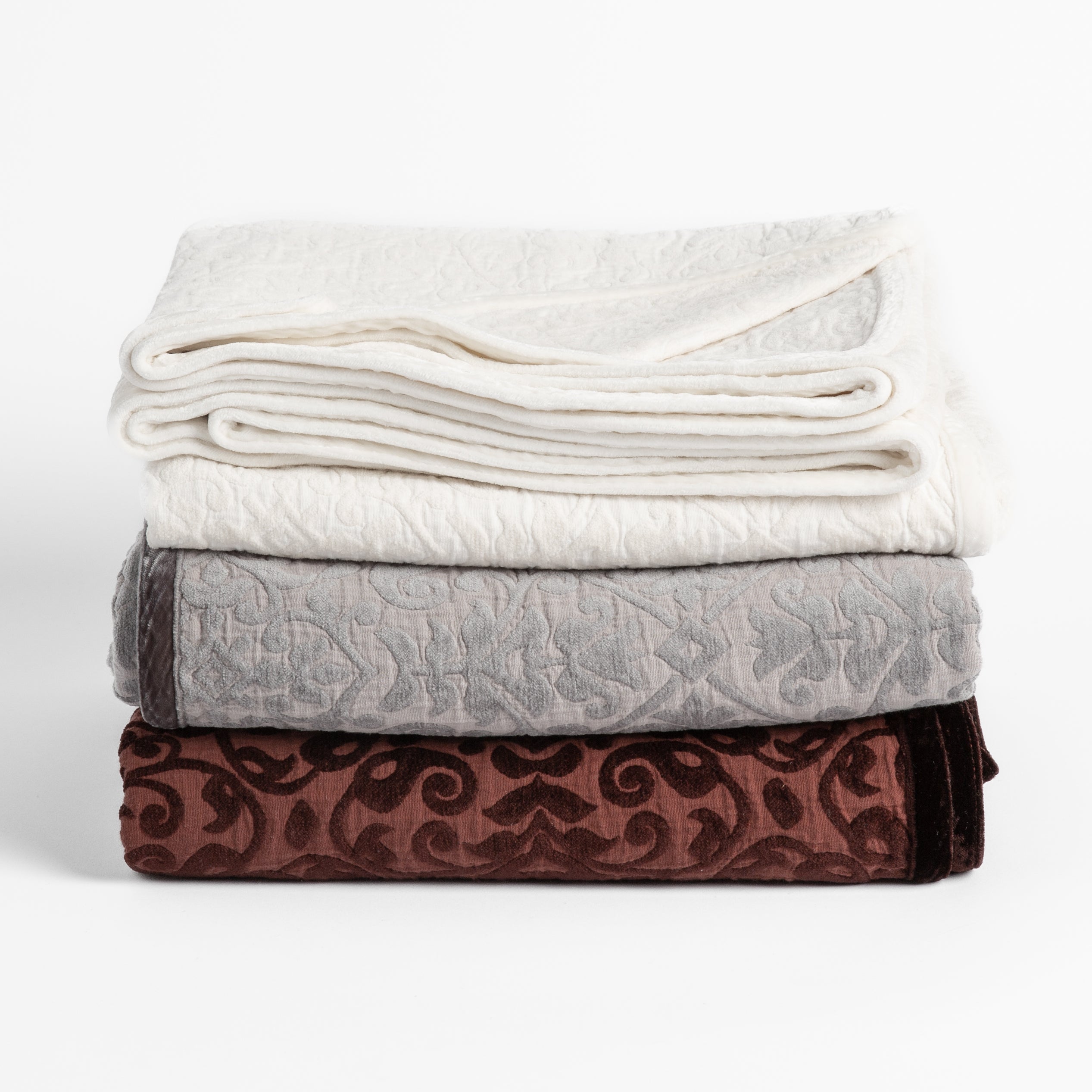 : three cotton jacquard chenile blankets in winter white, moonlight and mahogany folded and stacked neatly on a plain white background.