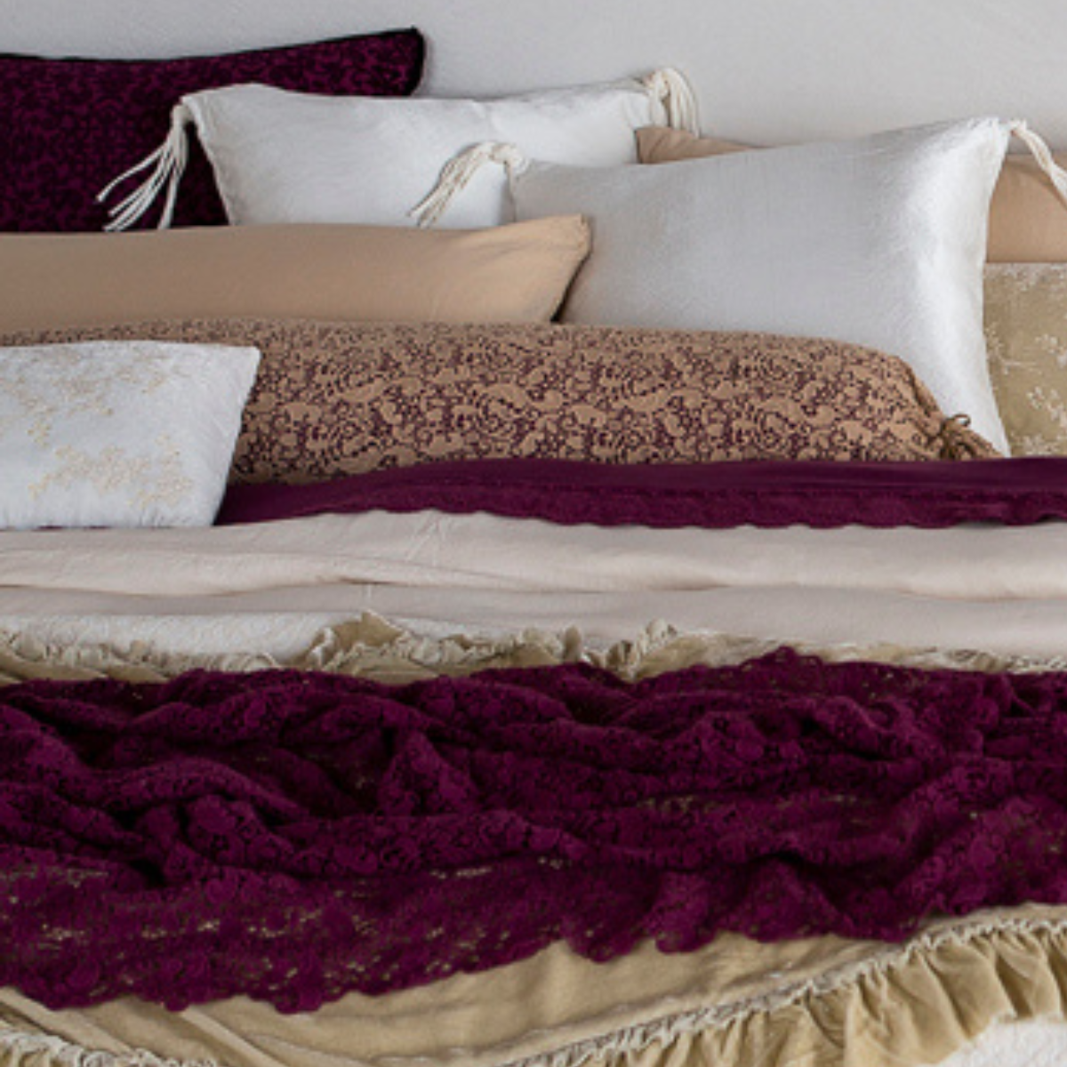 [allvariants]: a pulled in shot showing a lace bolster cover layered over a differently colored liner amongst a cluster of throw pillows, shams and sleeping pillows on a bed dressed in neutral tones with pops of the color fig. 
