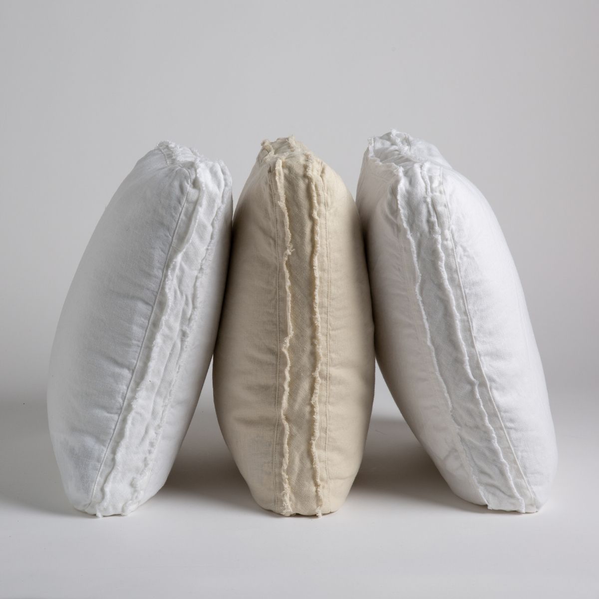 [allvariants]: three midweight linen throw pillows leaning on one another upright. Pillows shown in white, parchment and winter white. 