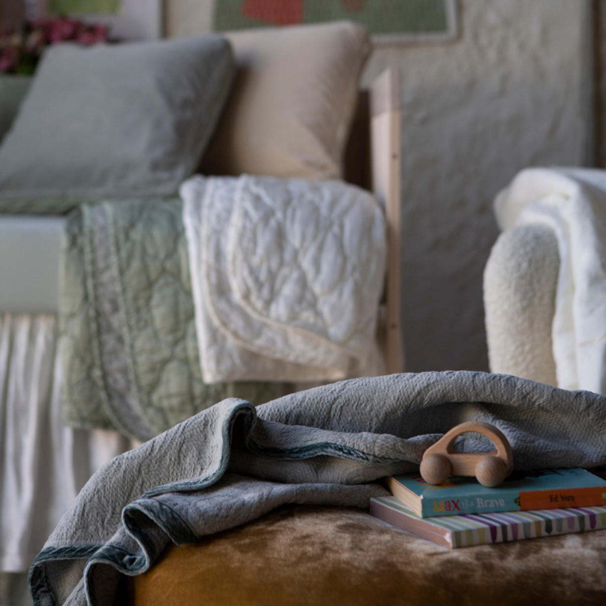 [allvariants]: Blanket in mineral draped over pouf, shown with books and toys in a green and cream toned nursery scene.