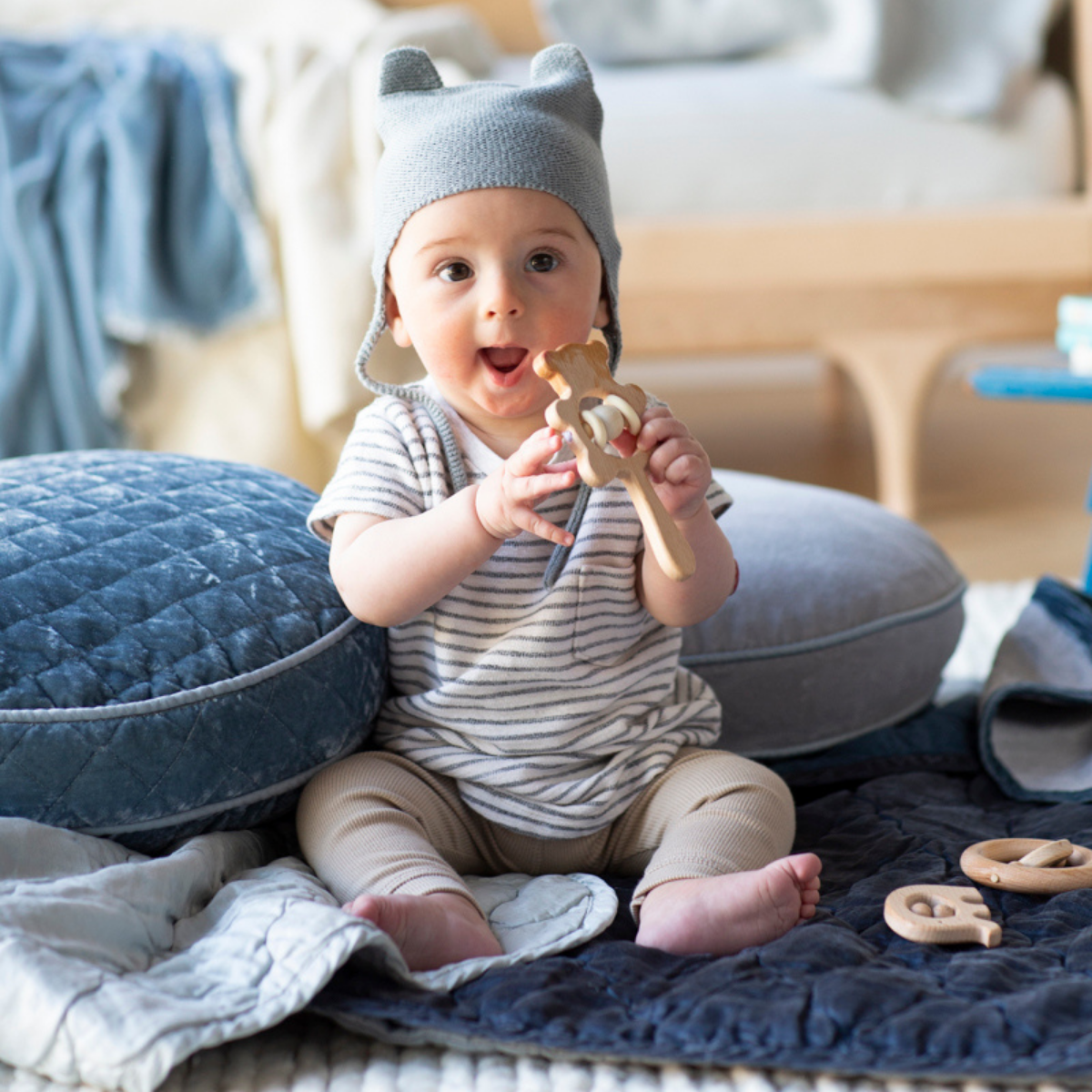 : Happy baby holding a wooden teether and sitting on rumpled blankets in cloud and midnight.