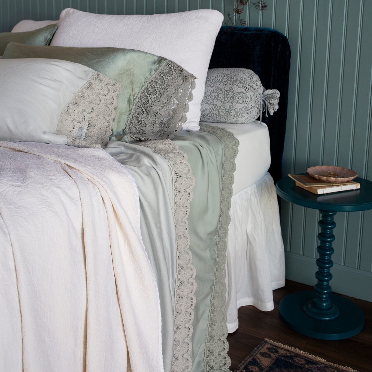 : two donella lace trimmed flat sheets from our Madera Luxe collection are shown in cloud and eucalyptus dressed with winter white fitted sheet, bedspread, shams and bed skirt. Lace trimmed pillowcases and a lace bolster are also shown at the head of the bed.  