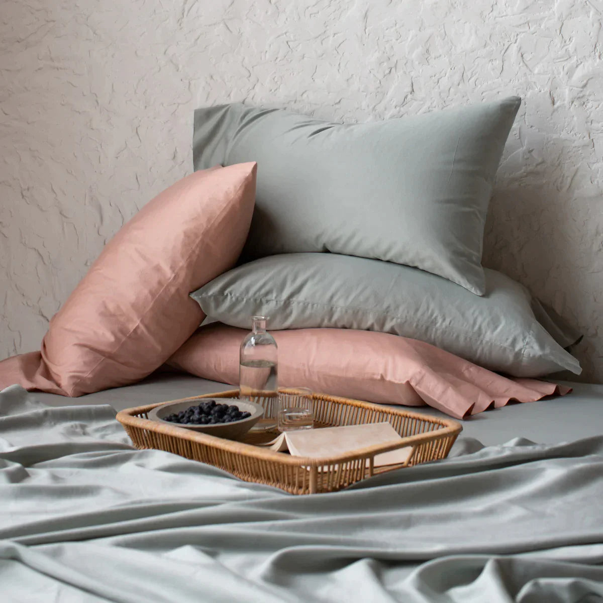 [allvariants]: Four Bria sleeping pillows in soft green and pink tones, stacked haphazardly on a matching bed against a textured white wall, perfect for lounging while enjoying the fruit and open book on the wicker tray.