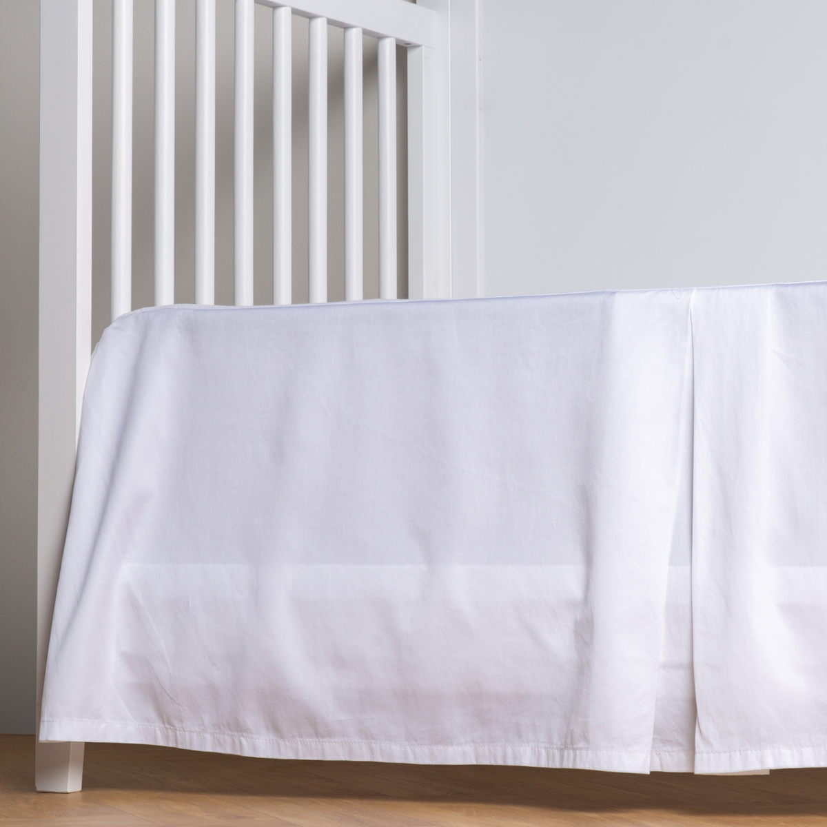 White: cotton sateen crib skirt with a center pleat shown straight on from a slight angle in a crib without a crib mattress. 
