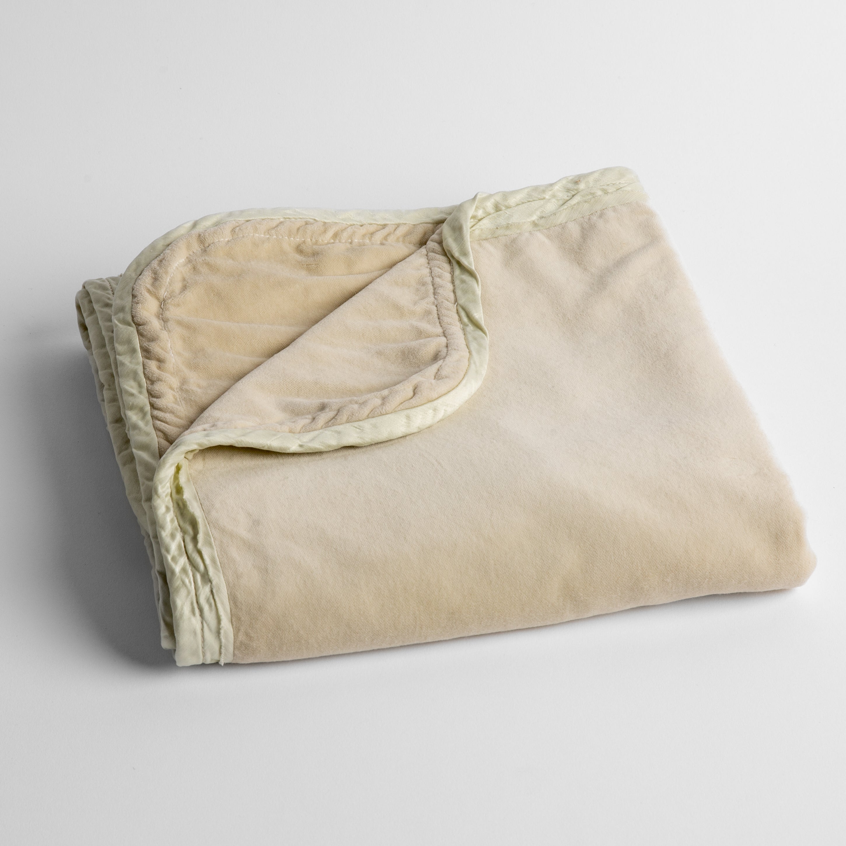 Parchment: cotton velvet baby blanket trimmed with charmeuse, folded with a covern folded back to show trim contrast; shot overhead and at a slight angle against a white background. 