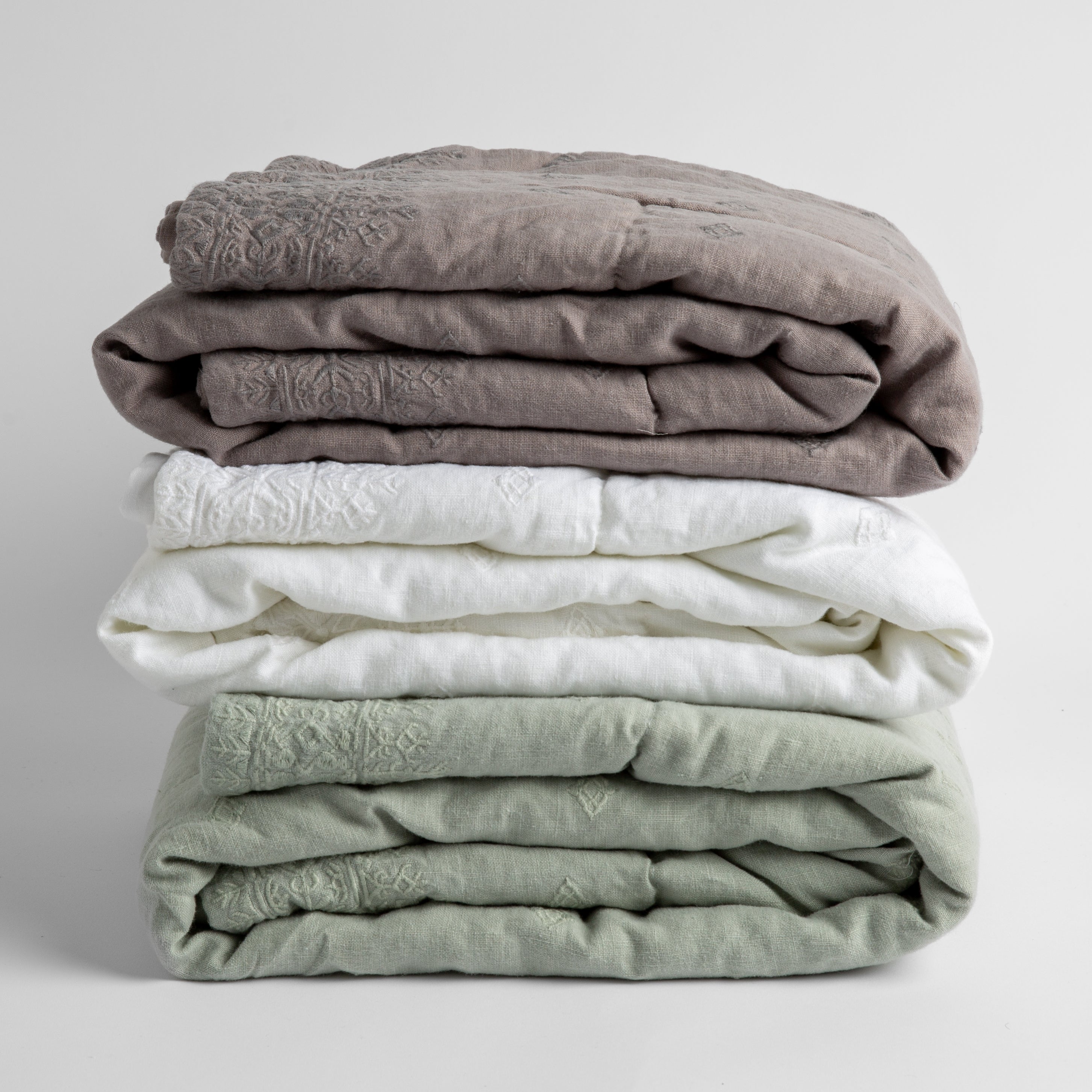 : A stack of three embroidered linen baby blankets in  moonlidhgt, white and eucalyptus.