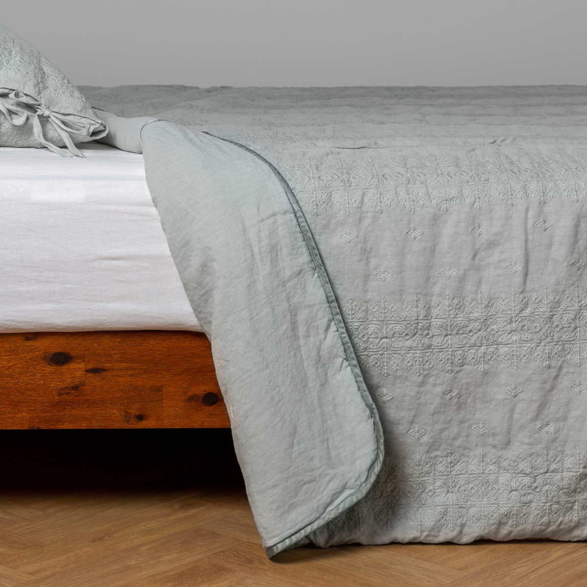 Eucalyptus: White: embroidered bedspread with corner pulled back  showing the plain linen back. Shown with a white fitted sheet and matching sham, the bedframe is slightly visible  and the bed is against a white wall and on a medium wood floor. 