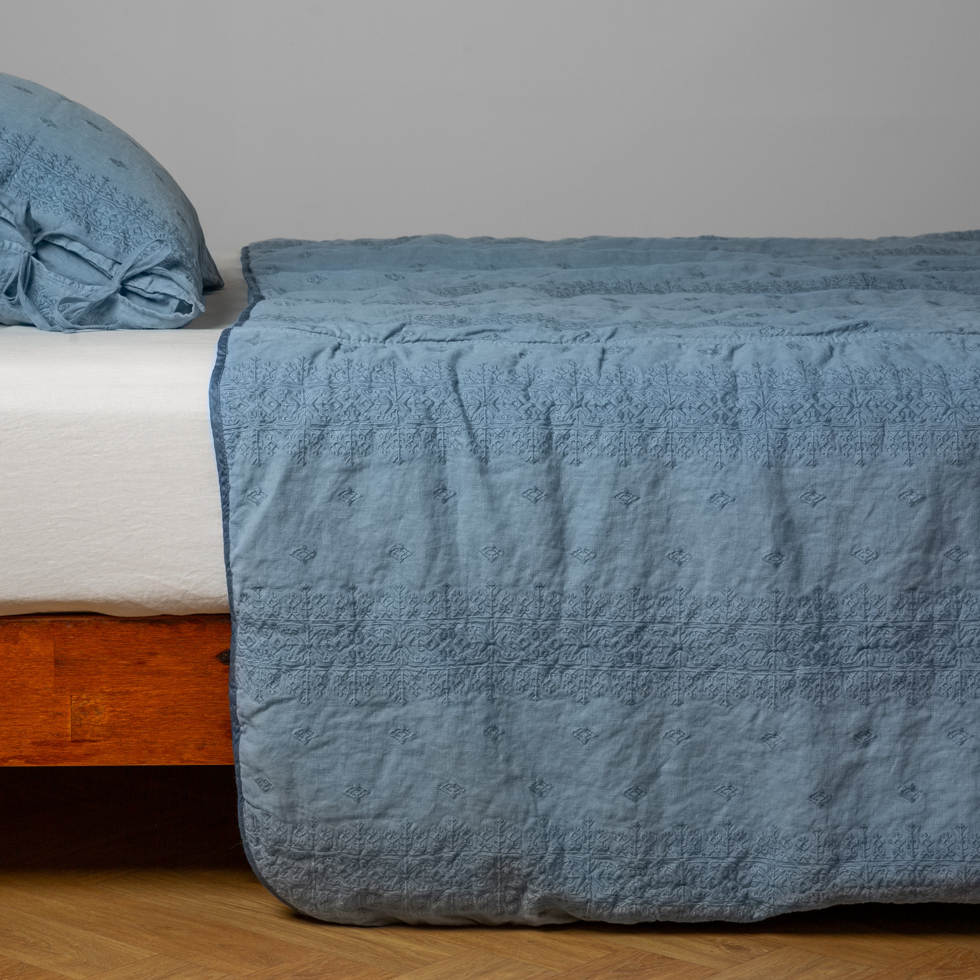 Tahoe Blue: embroidered midweight linen bedspread with matching sham on a bed with a white fitted sheet. Shot from the side view, the bedframe is slightly visible  against a white background and medium wood floor. 