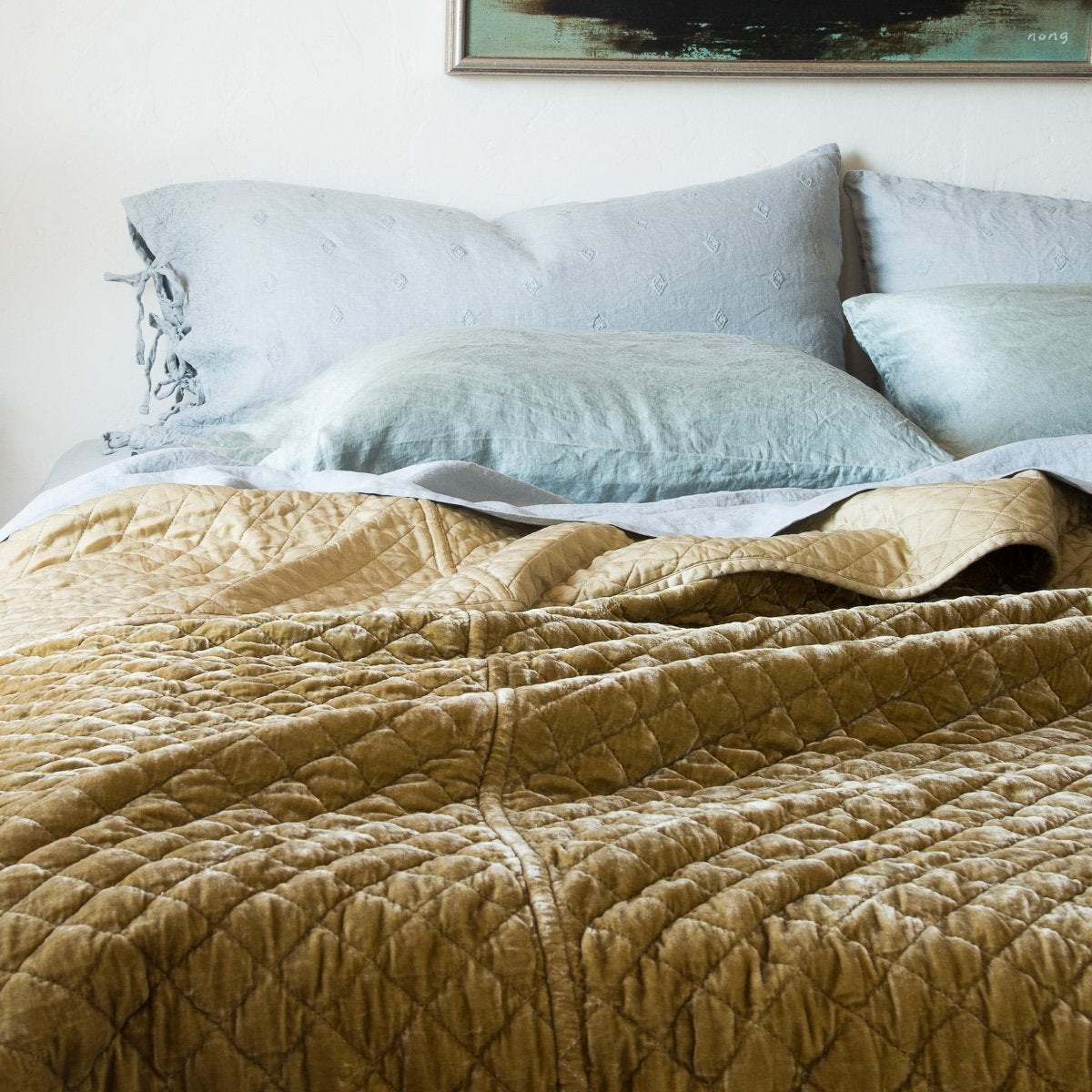 : Ines standard shams in cloud, layered behind on-tone linen sheets and silk charmeuse sleeping pillows, and a rumpled silk velvet quilted coverlet in a rich golden tone - cropped head-on view.