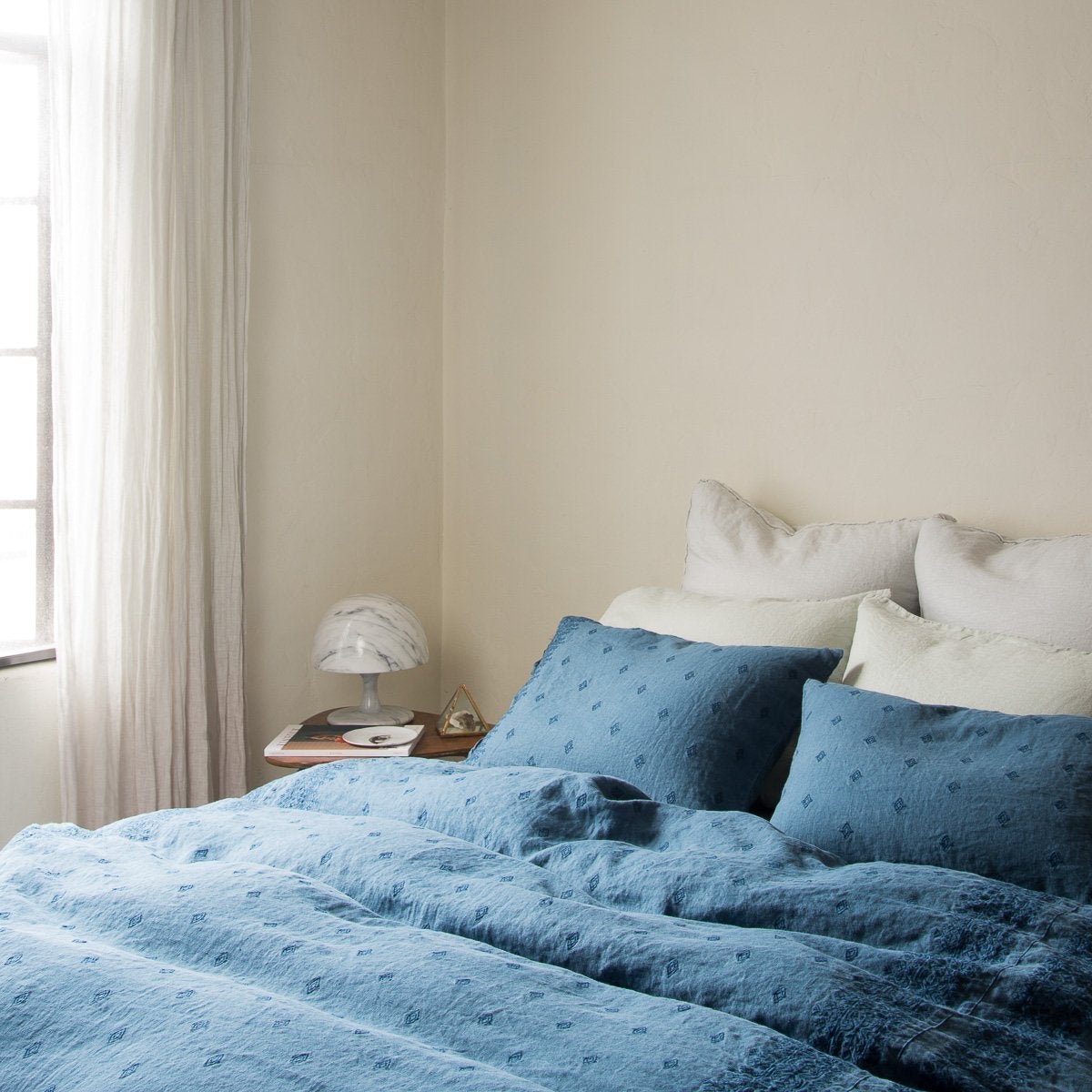 : Midnight Ines shams and duvet cover, layered with soft green linen pillows, with light toned walls and curtains, all bathed in natural light - cropped three-quarter angle.