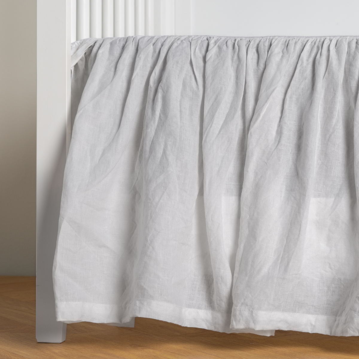 White: crib skirt shown on a white crib with no mattress against a white wall and medium wood flooring. 