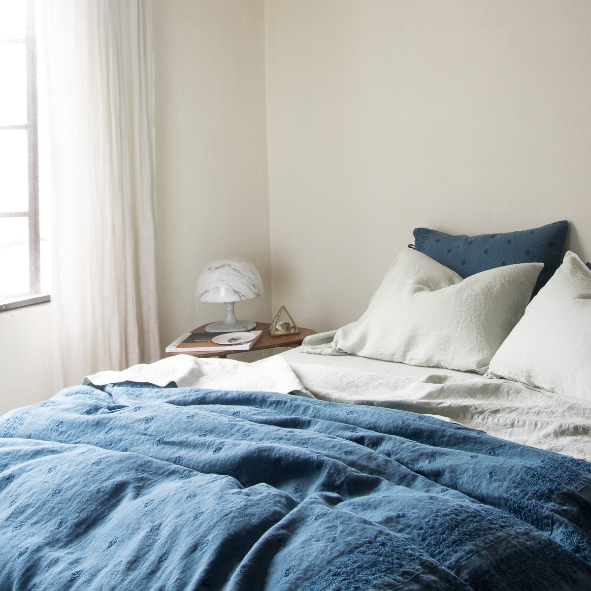 [allvariants]: Linen pillowcases in eucalyptus with matching sheets with a rumpled duvet in midnight blue embroidered linen - cropped three-quarter angle.
