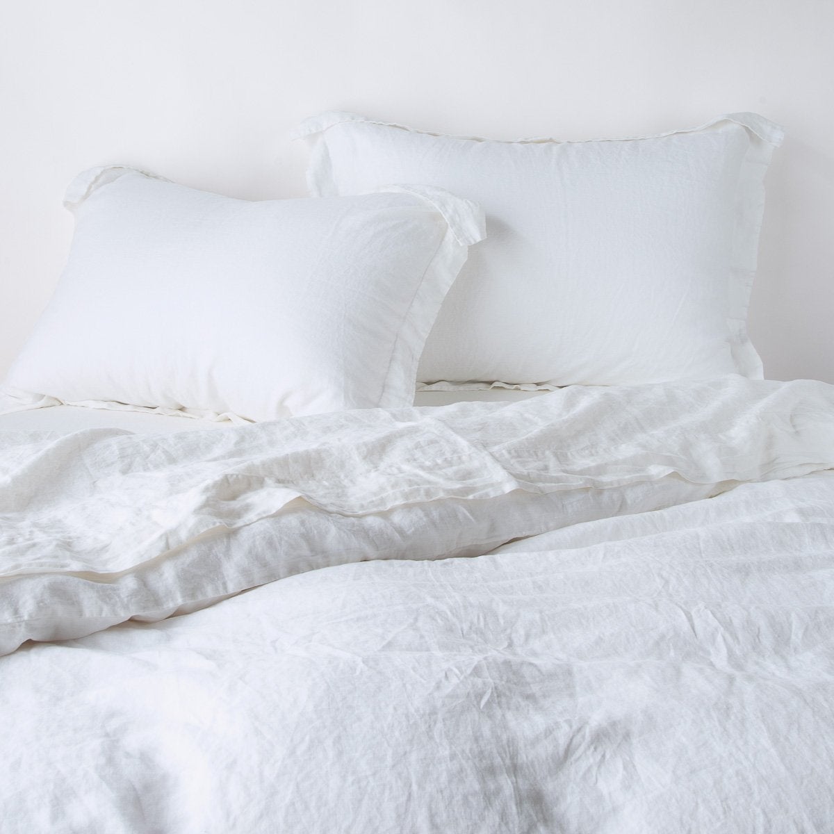 White: Two shams leaning upright against a white wall with matching sheeting and duvet - cropped end of bed view.