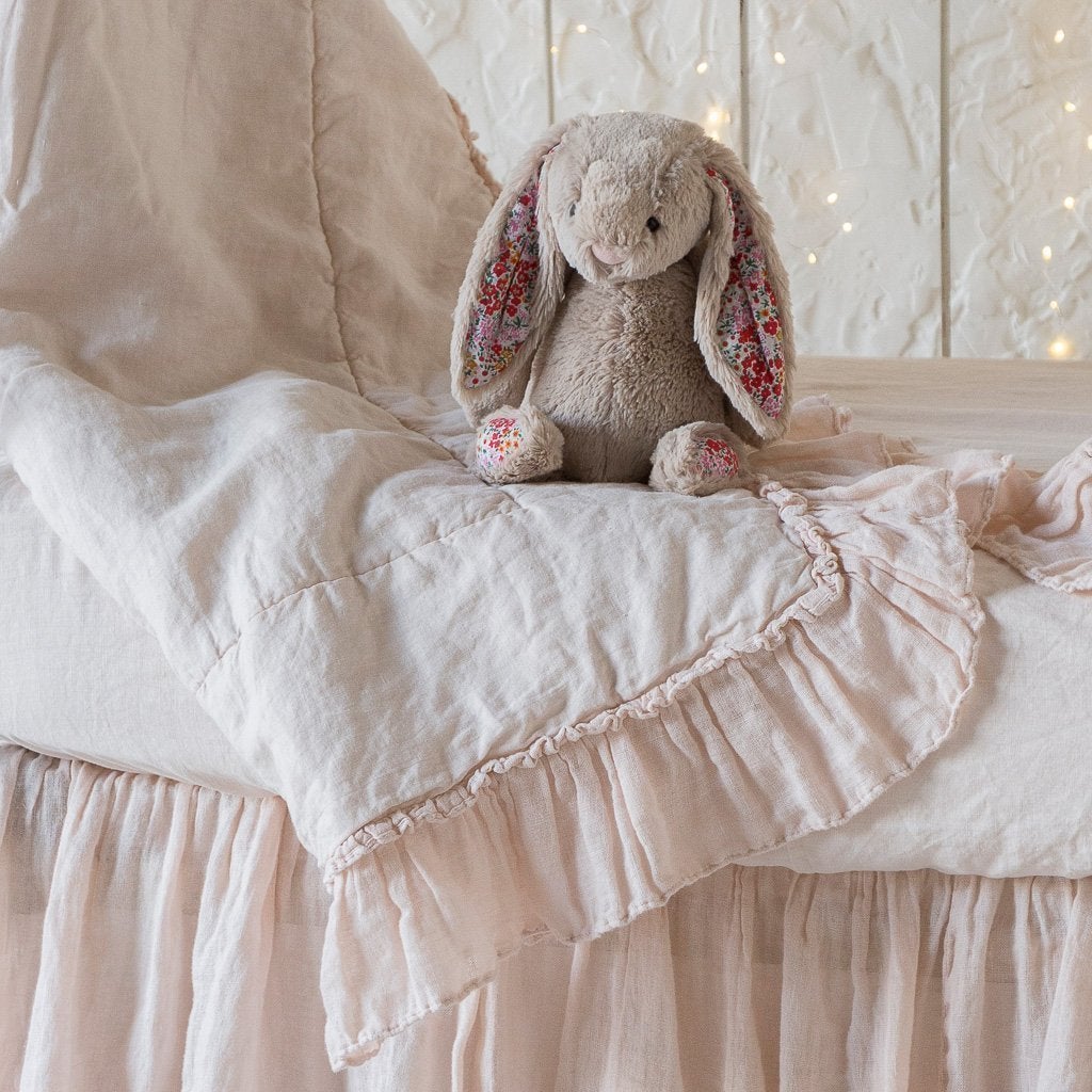 : Linen whisper baby comforter in pearl, draped over matching sheet and crib skirt, shown with a stuffed bunny and twinkly lights - side view.