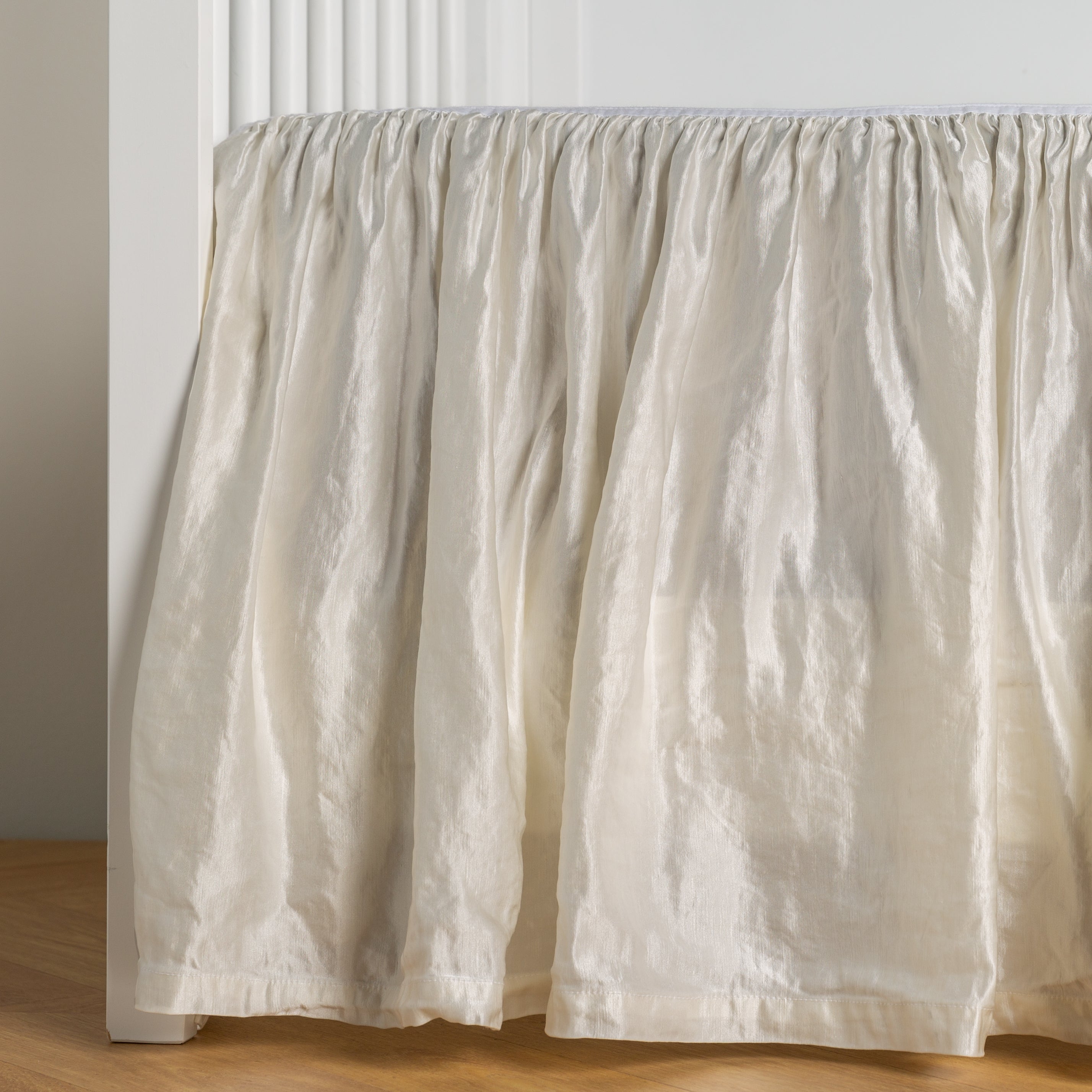 Winter White:  charmeuse crib skirt shown on a white crib against a white wall and medium wood floor. 