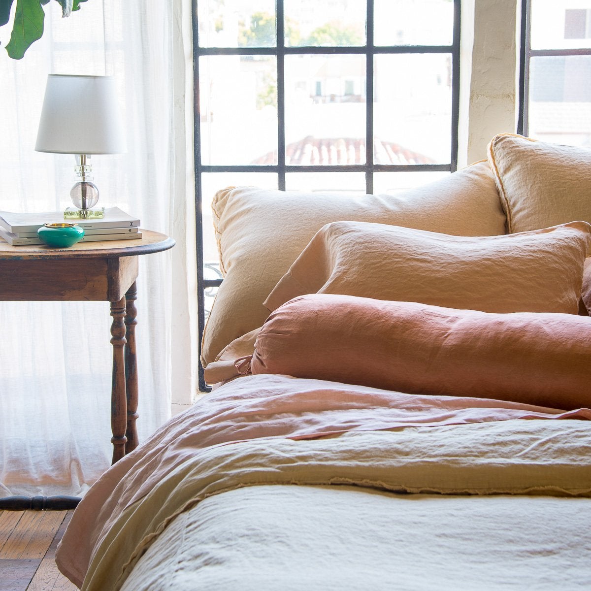 [allvariants]: Paloma sleeping pillow and bolster on linen bedding, in soft pink and gold tones against big windows - cropped end of bed view.