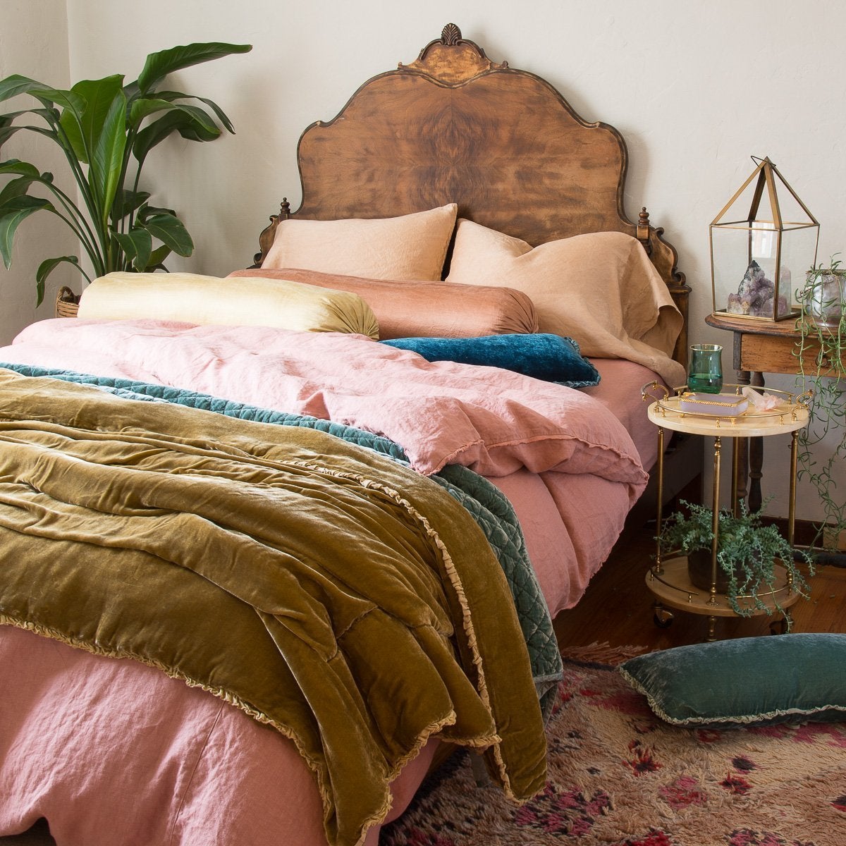 : Paloma sleeping pillows and bolsters layered with linen and silk velvet, in pink and gold with pops of blue and green. Three-quarter angle showing dramatic wooden headboard and tall greenery.