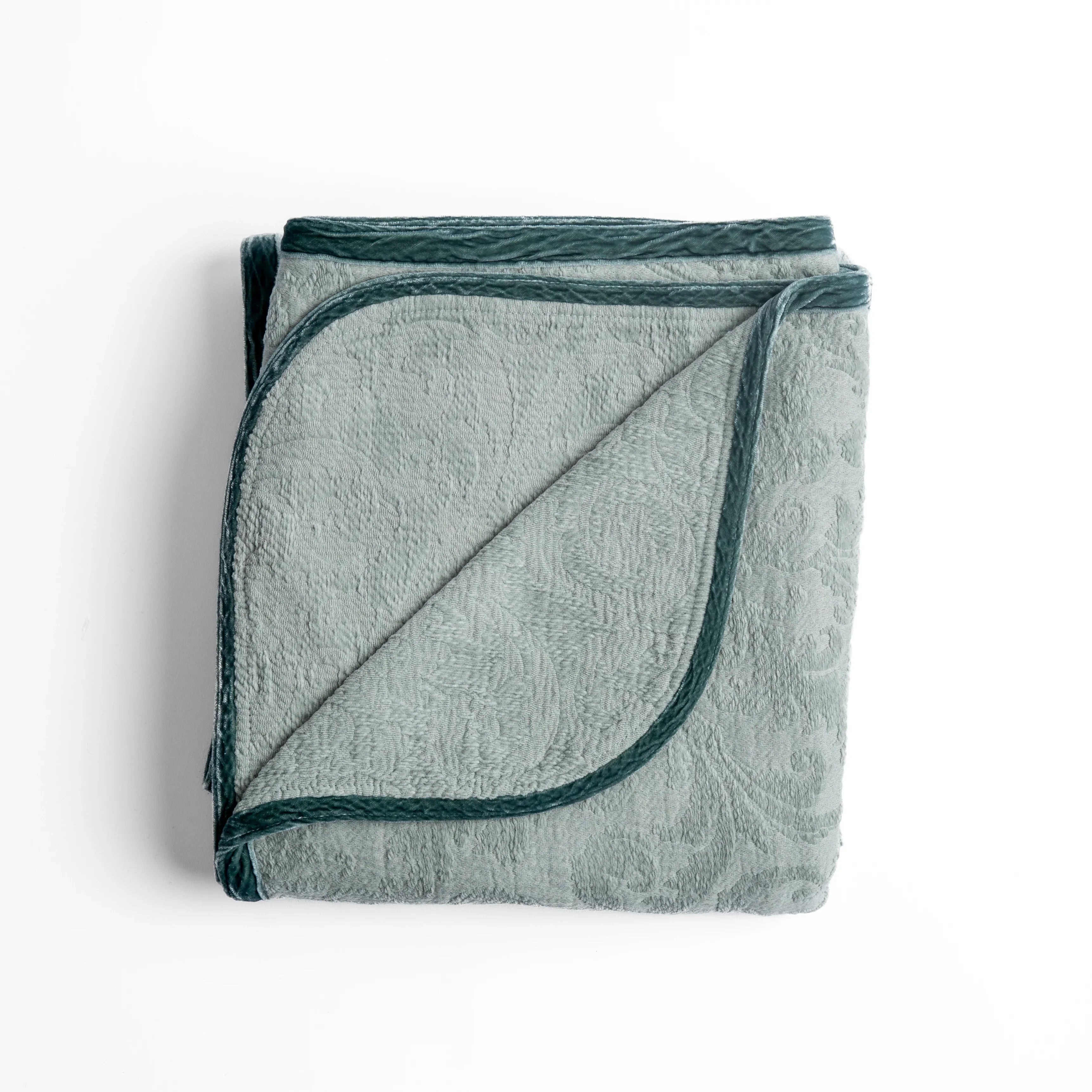 Eucalyptus: folded organic cotton damask throw blanket shown with the corner folded down to show silk velvet trim detail — overhead against a plain white background. 