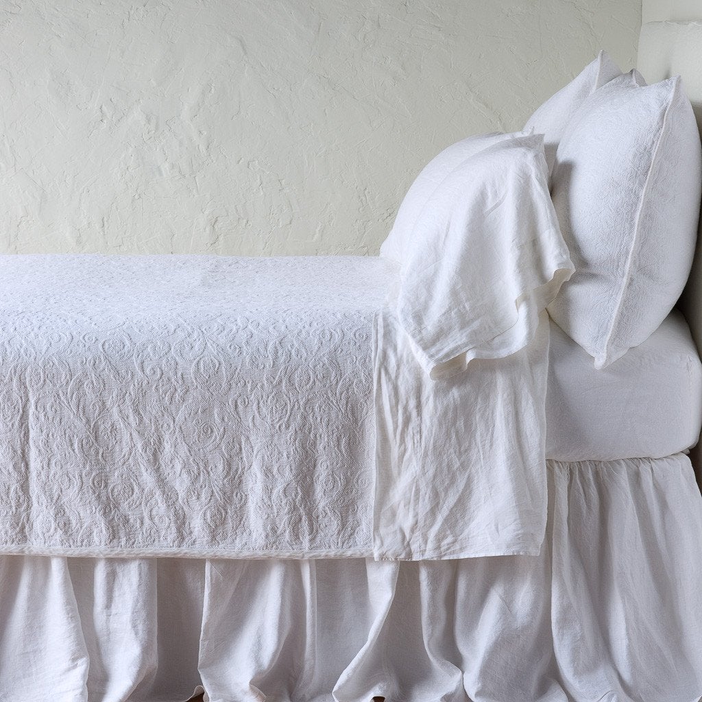 White: The Adele coverlet in white, on a bed viewed from the side against a plain white wall. The bed is styled neatly with the coverlet folded back to reveal white sheets, bed skirt and pillows.