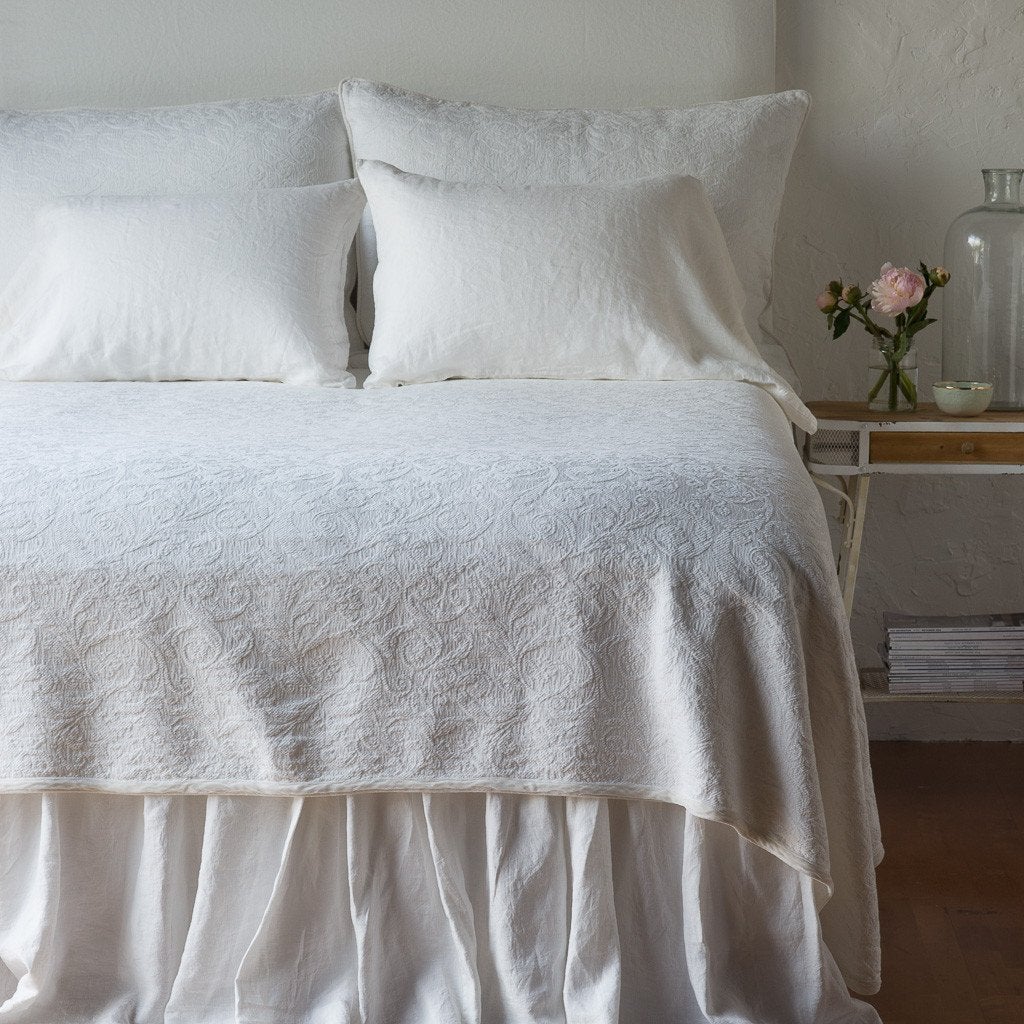 Winter White: The Adele coverlet in winter white, viewed from the foot of the bed, with winter white bed skirt and pillows visible. The headboard is plain light color, and part of a bedside table is visible with flowers, a vase and some books. against a plain white wall.