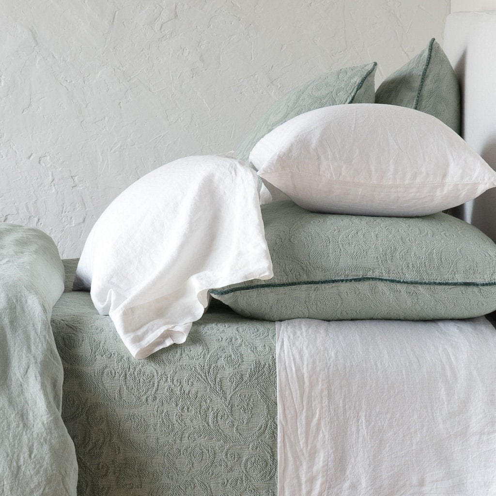 [allvariants]: Adele shams in eucalyptus on a bed shown from the side, layered with white sheets and pillowcases and a eucalyptus coverlet against a plain white wall.