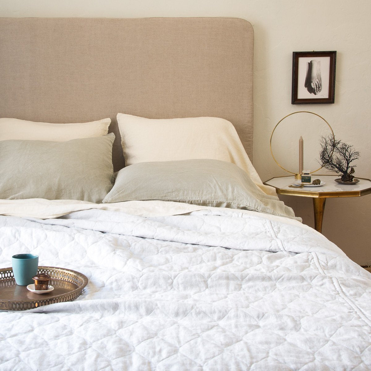 [allvariants]: White Austin quilted coverlet shown partially unmade from the foot of the bed with light neurtal sheets and pillowcases, against a beige headboard. Styled with a tray on the bed and simple bedside table and art.